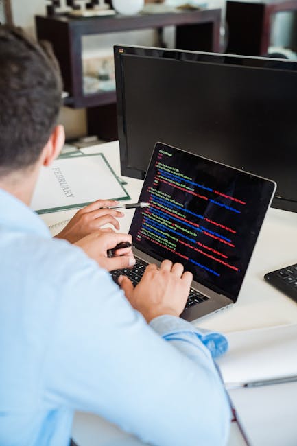 A person with short dark hair, dressed in a light blue shirt, is seated at a desk working on a laptop that displays colorful lines of computer code. The workspace includes a black computer monitor, a 