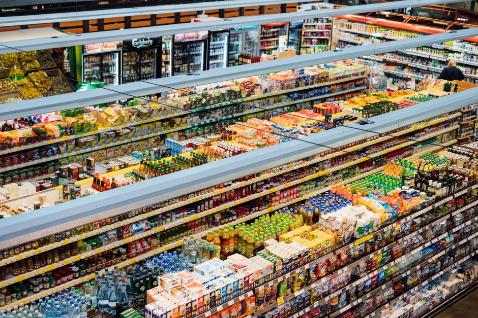 An extensive view of supermarket shelves in a retail shop after refurbishment, showcasing neatly arranged rows of bottled beverages, snacks, and packaged goods. The shelves are made of light-colored m