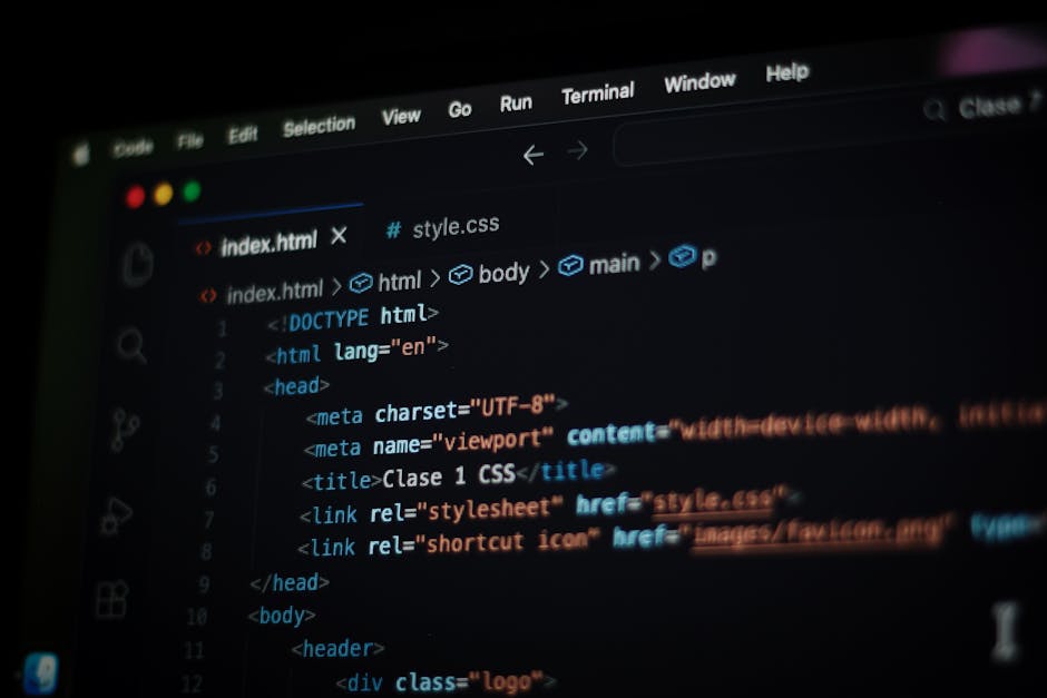 A person with short dark hair, wearing a light blue shirt, working on a laptop displaying colourful lines of code. The workspace includes a desk with a closed magazine titled 'FURNITURE', a black comp